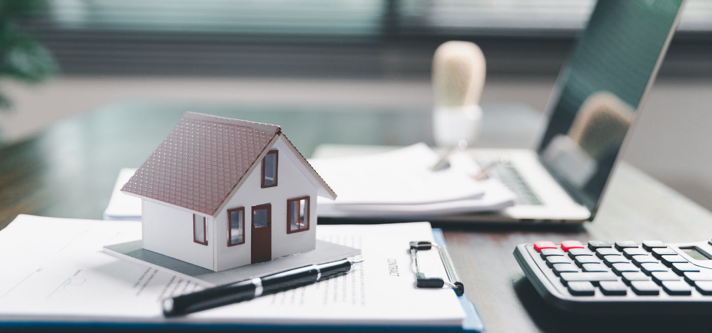 Casa em miniatura sobre documentos, com calculadora e portátil ao fundo